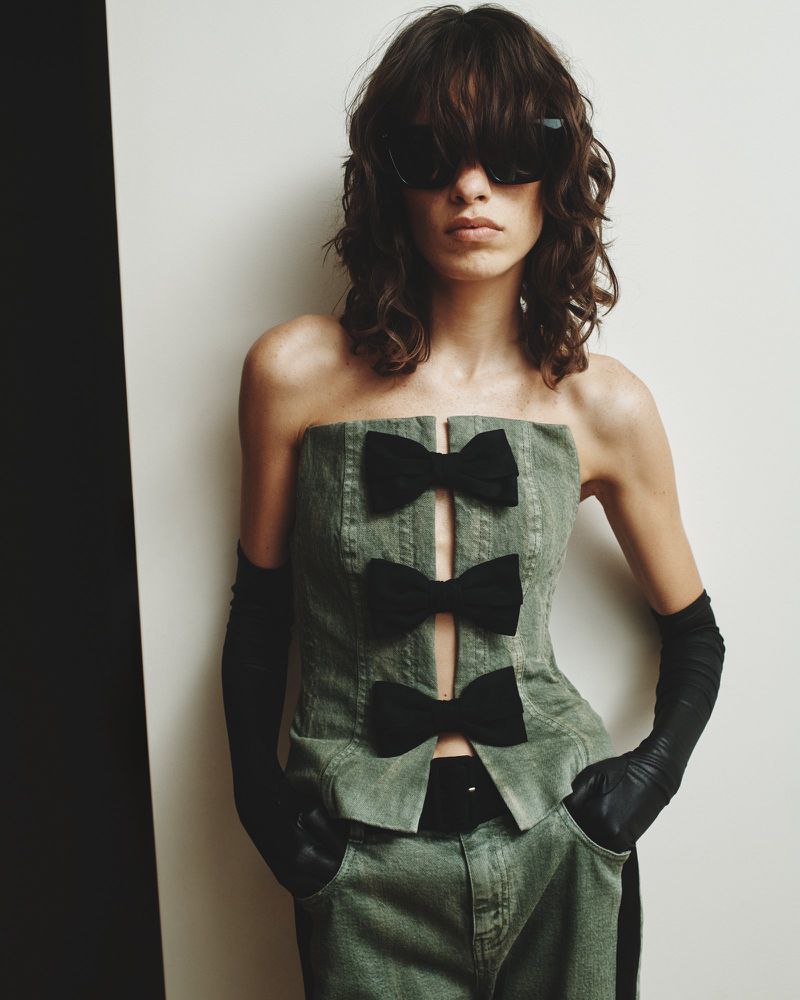 A woman wearing a strapless green denim top with black bows, matching pants, gloves, and sunglasses poses against a neutral backdrop.