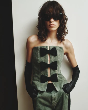 A woman wearing a strapless green denim top with black bows, matching pants, gloves, and sunglasses poses against a neutral backdrop.