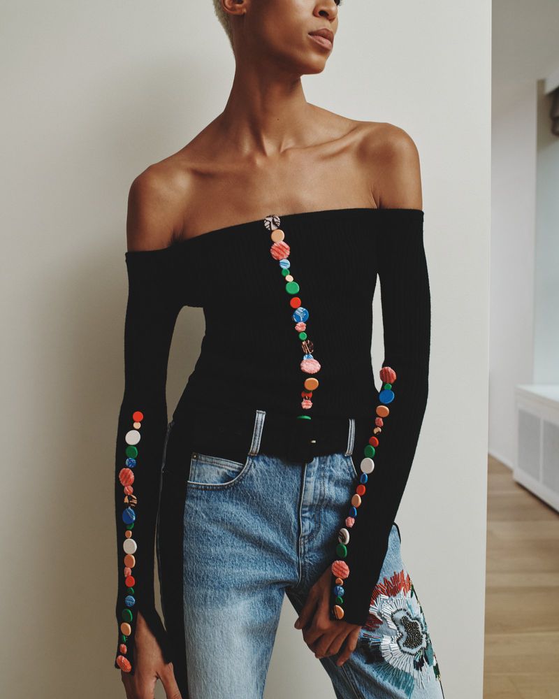 A woman models a black off-the-shoulder top with colorful button accents paired with blue jeans with floral embroidery in a studio setting.