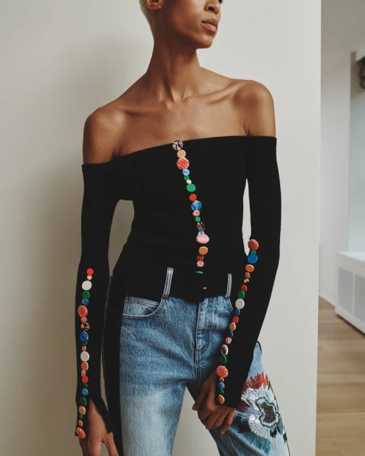 A woman models a black off-the-shoulder top with colorful button accents paired with blue jeans with floral embroidery in a studio setting.