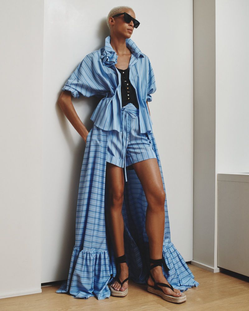 Woman in blue and white striped shorts suit with a long flowing train, accessorized with black sandals and sunglasses, poses indoors.