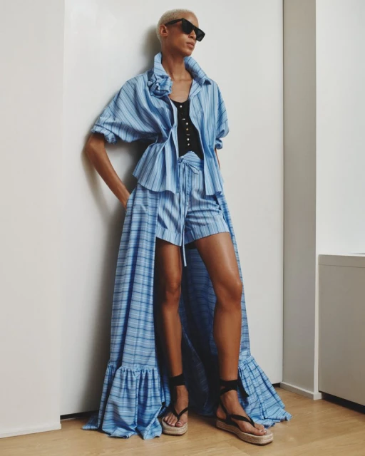 Woman in blue and white striped shorts suit with a long flowing train, accessorized with black sandals and sunglasses, poses indoors.