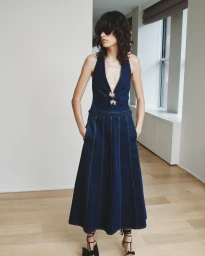 Model wearing a deep V-neck denim halter dress with decorative buttons in a bright interior room for Hellessy SS25.