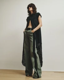 Model standing against a light background wearing a dark longline shirt and green wide-leg trousers for Hellessy SS25.