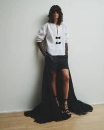 Model standing against a white wall wearing a white blouse with black bows and a long black tulle skirt overlay