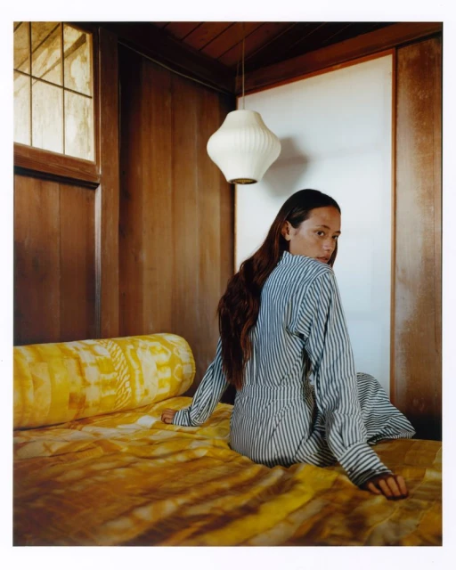 Brunette woman in striped shirt dress sits on a yellow patterned bed looking over her shoulder in a wood-paneled room with natural light.