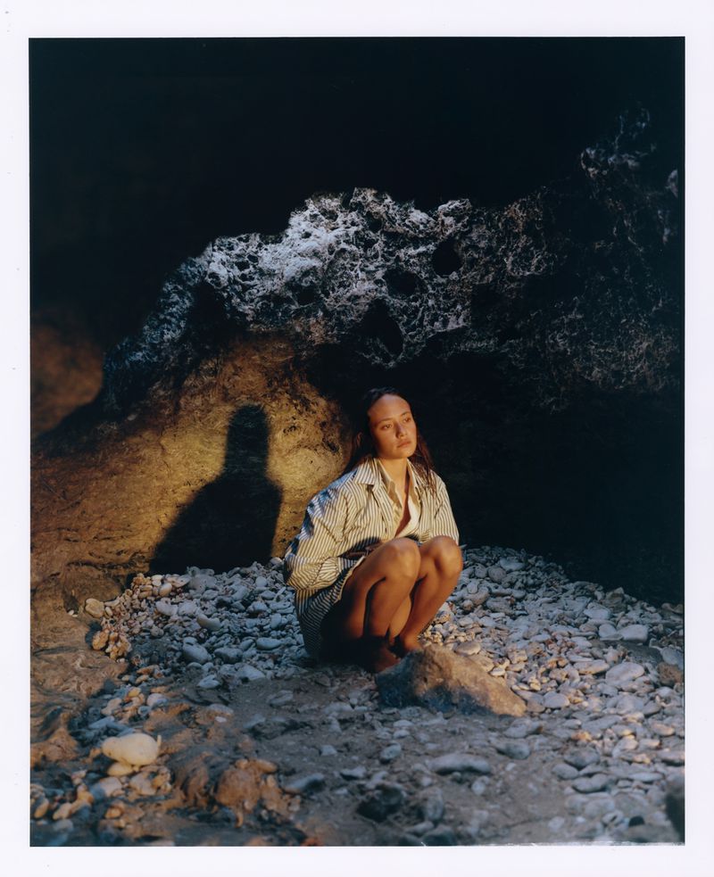 Young woman crouches in a rocky cave with a neutral expression, lit by warm light against a dark background, wearing a striped shirt.