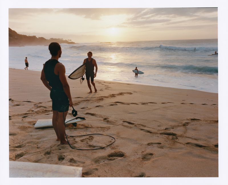 Seascape captures three surfers at sunset: one walks with board, one stands, and another enters the water with their surfboard.