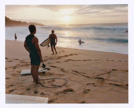 Seascape captures three surfers at sunset: one walks with board, one stands, and another enters the water with their surfboard.