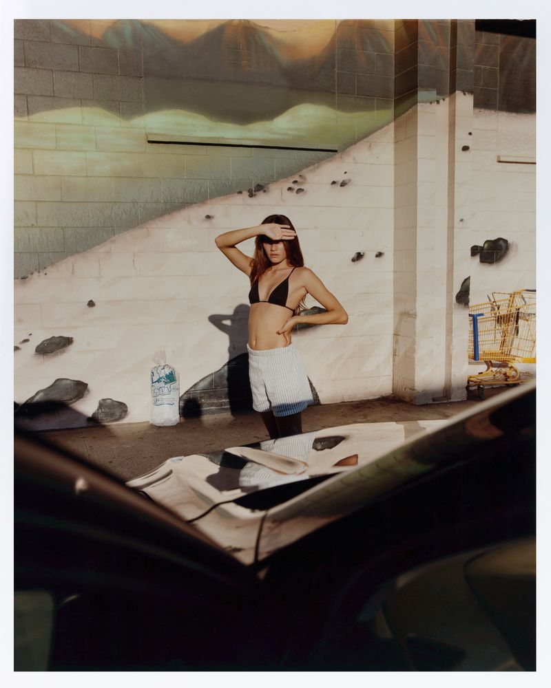 Woman in bikini top and shorts shields eyes in an urban setting near a car, with a painted mural wall in the background.