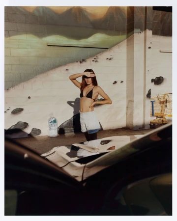 Woman in bikini top and shorts shields eyes in an urban setting near a car, with a painted mural wall in the background.