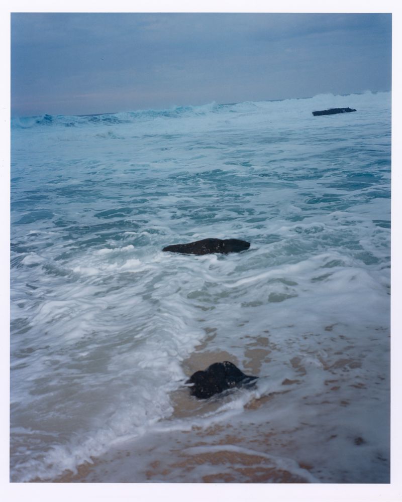 Ocean waves crashing onto a sandy beach with dark rocks and a cloudy sky above in an eye-level, outdoor scenic shot.