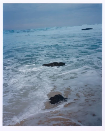 Ocean waves crashing onto a sandy beach with dark rocks and a cloudy sky above in an eye-level, outdoor scenic shot.