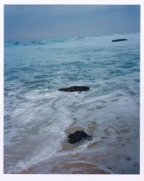 Ocean waves crashing over dark rocks on a sandy beach under an overcast sky.