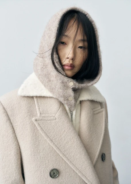 Asian female model in a beige wool coat with a hooded collar posing in front of a neutral background in a fashion editorial shot.