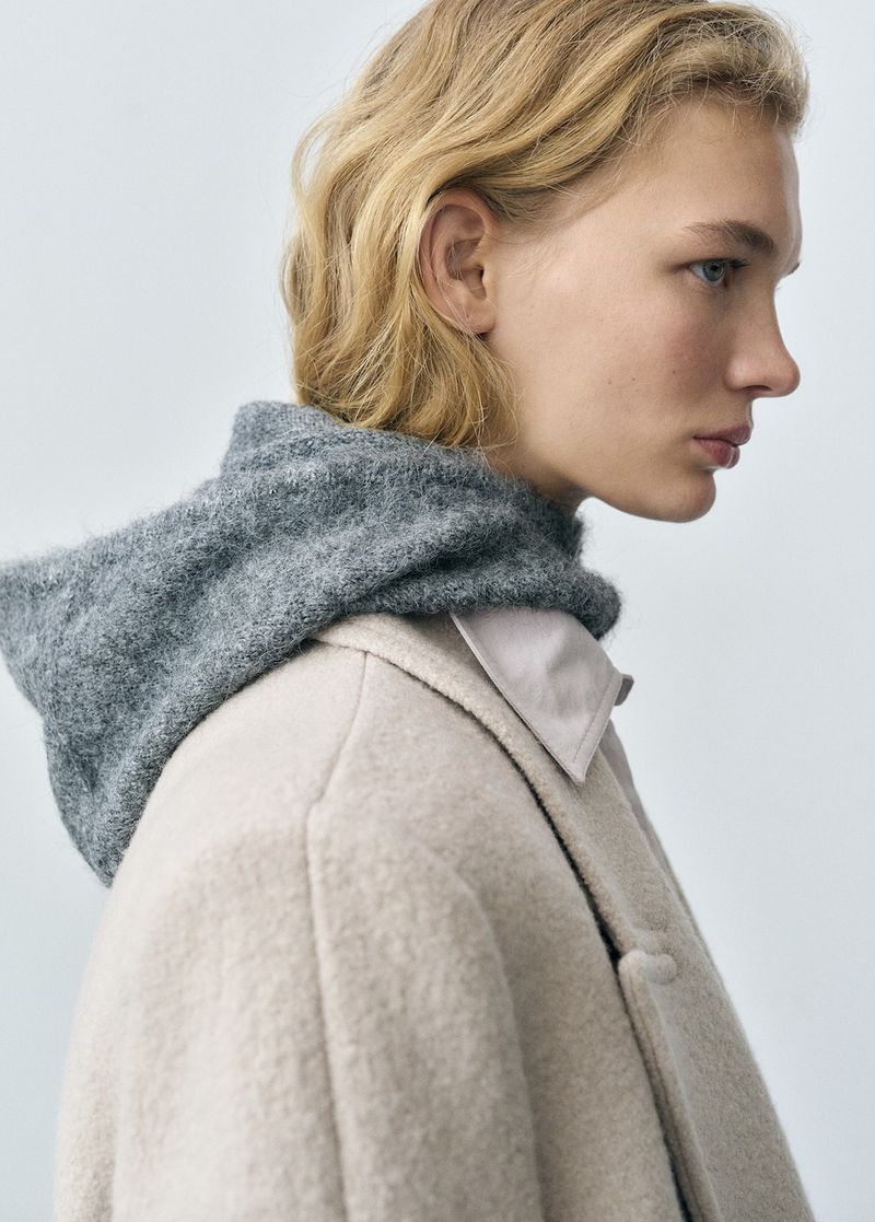 Fashion editorial shot: a young fair-skinned woman with blonde wavy hair, wearing a wool coat and grey hooded sweater.