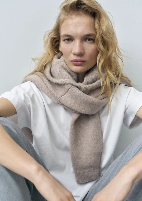 Portrait of a woman with blonde curly hair wearing a beige scarf and a white t-shirt posing in a photo studio setting.