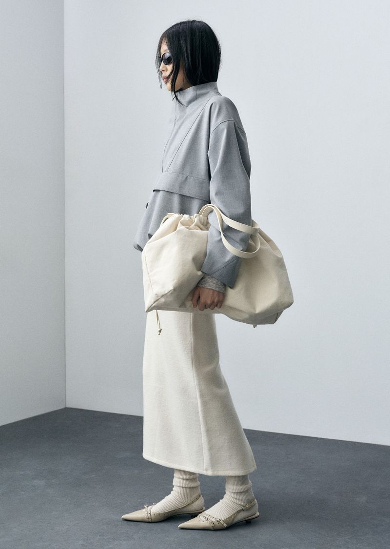 Woman in grey top, cream skirt, and ballet flats carries a large, light-colored drawstring bag in a minimalist studio setting.
