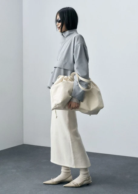 Woman in grey top, cream skirt, and ballet flats carries a large, light-colored drawstring bag in a minimalist studio setting.