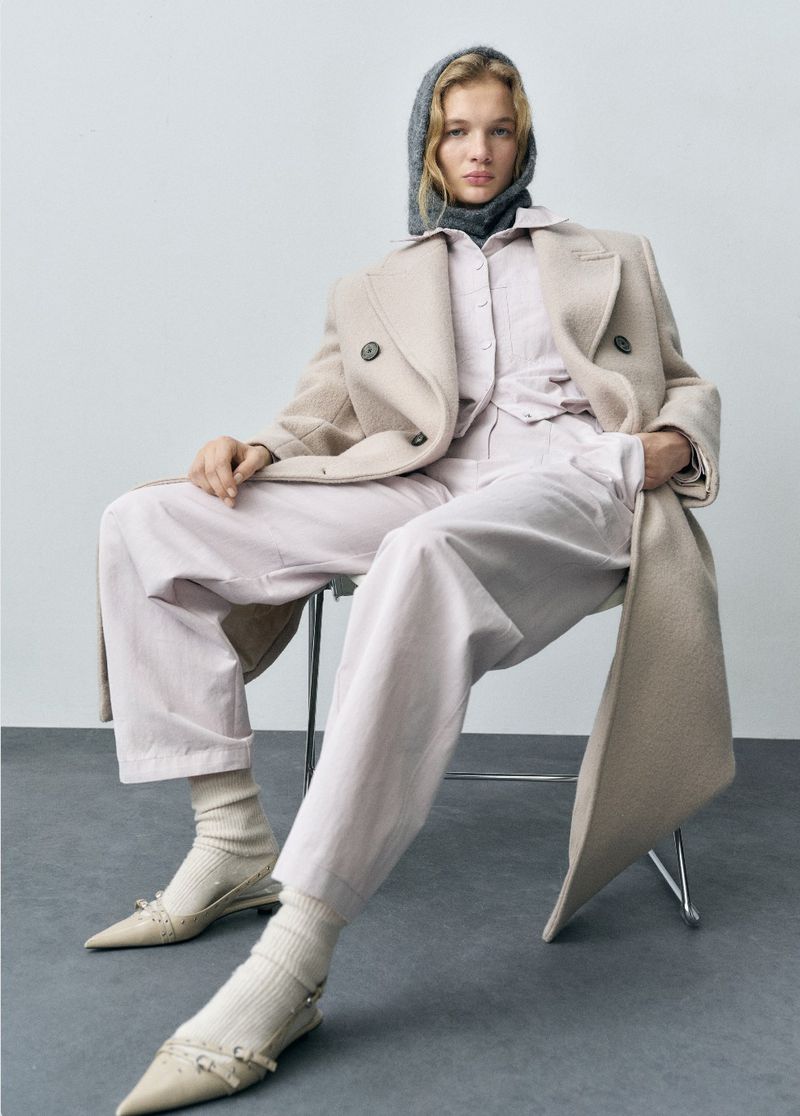 Fashionable woman sits on chair in light pink linen set with beige coat, socks, and neutral pointed shoes against light gray studio backdrop.