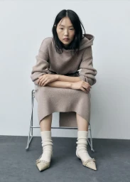 Model seated on a metal chair wearing a beige hooded knit dress, cream socks, and flats against a grey backdrop