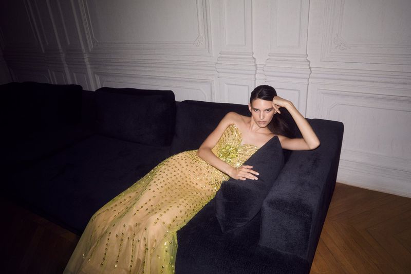 Elegant adult female poses in a strapless yellow embellished gown on a velvet couch in an indoor setting with a white wall backdrop.