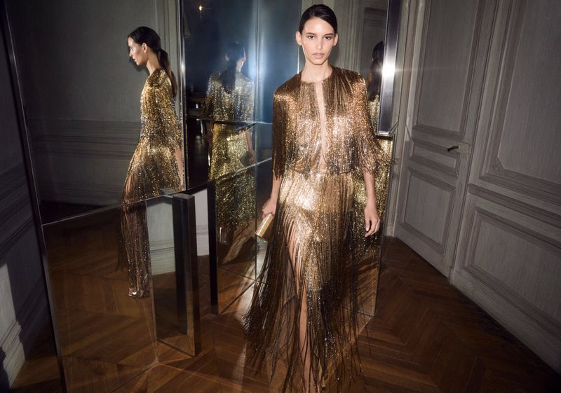 Fashion model in a shiny gold fringe dress holds a gold clutch, posing in a room with mirrors and gray-paneled walls.