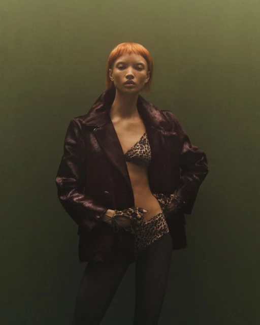 Elegant adult woman in a leopard-print bikini and maroon jacket poses indoors against a green backdrop, showcasing a high-fashion style.