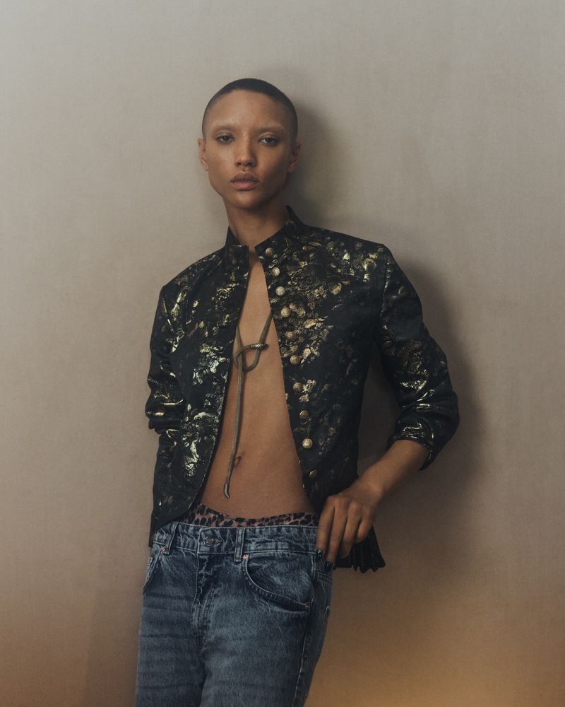 A slim adult female poses in a black jacket, blue jeans, and leopard-print underwear in front of a beige wall. Fashion shot, indoors.