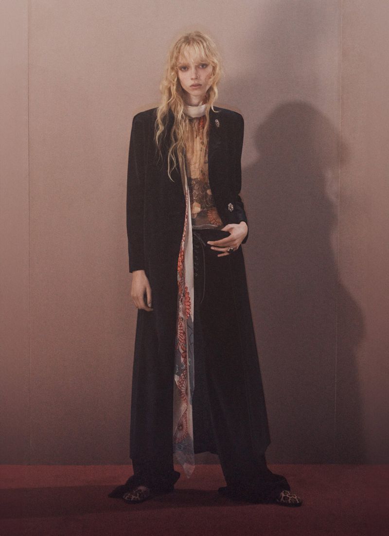 A solitary fair-skinned female fashion model stands in a studio setting wearing a long black coat, wide-leg pants, and a patterned scarf.