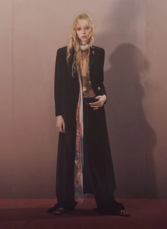 A solitary fair-skinned female fashion model stands in a studio setting wearing a long black coat, wide-leg pants, and a patterned scarf.