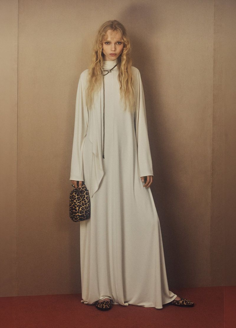 A fair-skinned young woman with blonde wavy hair stands in a long-sleeved white dress, holding a leopard-print bag. She faces the camera.