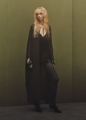 Model with long blonde hair wearing a black dress and dark coat standing against a muted olive green studio backdrop