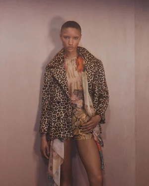 Model with a shaved head wearing a leopard print coat and snakeskin print shorts against a plain neutral wall