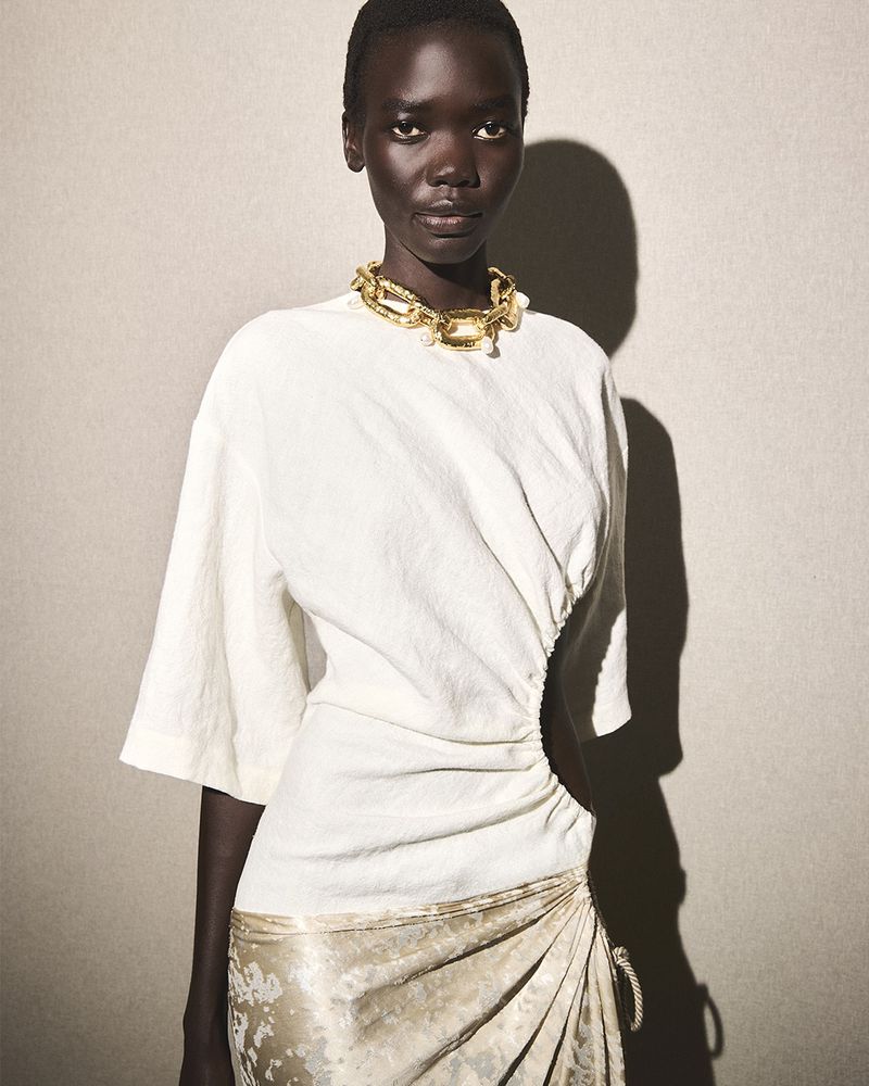 Close-up fashion shot of a dark-skinned woman in a white top and gold skirt, featuring a gold chain necklace and neutral backdrop.