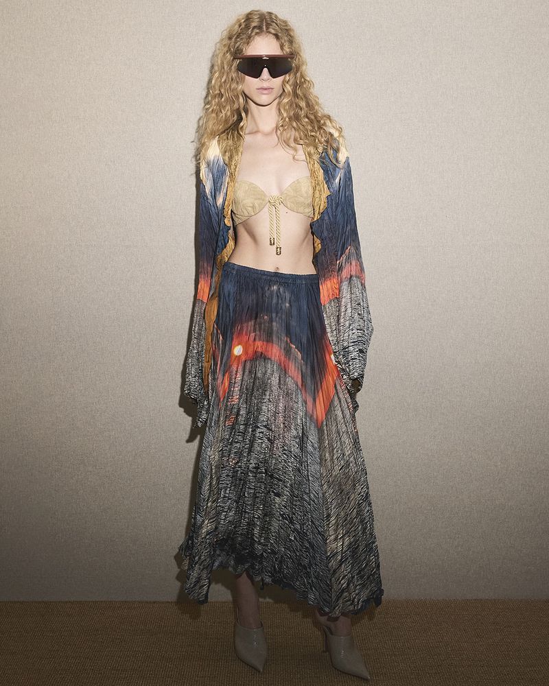 A young adult woman with curly blond hair poses indoors wearing sunglasses, a bra top, a long skirt, and a cardigan with a desert landscape print.