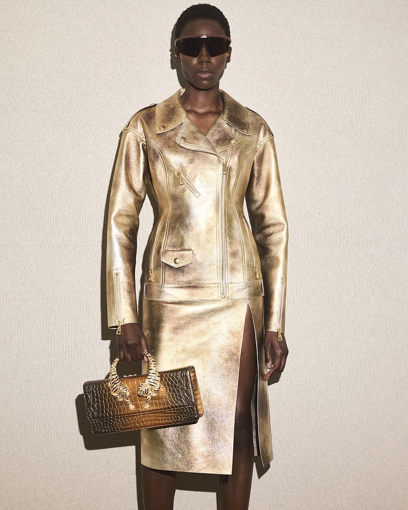 Woman in gold jacket and skirt with handbag. Dark-skinned female model wearing sunglasses and a gold leather outfit poses in studio.