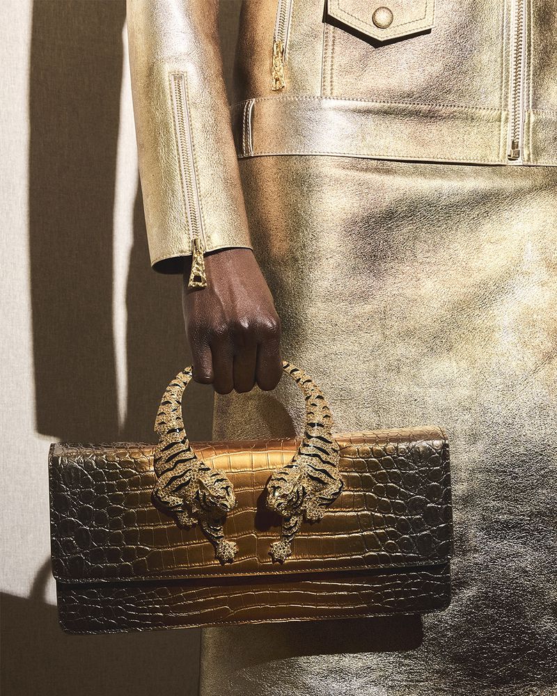 Gold-toned handbag with tiger-shaped handles held by a dark-skinned hand; model wears gold jacket and skirt, studio lighting.