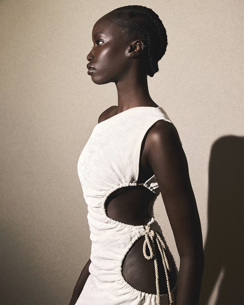 Side profile studio shot of a Black woman with braided hair wearing a cream-colored cutout dress with rope ties against a neutral backdrop.