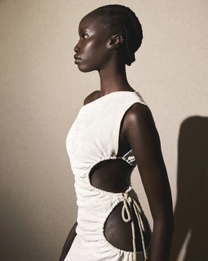 Model in profile wearing a white one-shoulder cut-out dress against a neutral textured studio background.