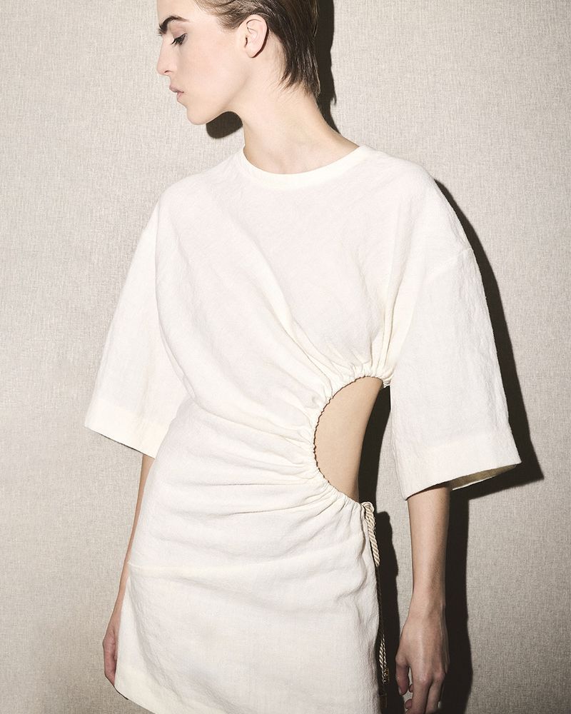 Model wearing a white cut-out dress against a textured neutral background with a sharp side shadow.