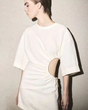 Model wearing a white cut-out dress against a textured neutral background with a sharp side shadow.