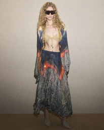 Model with long curly blonde hair wearing dark sunglasses, patterned long-sleeve robe, and maxi skirt against a neutral textured wall.