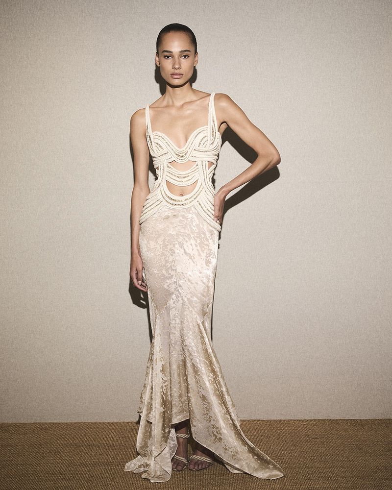Model wearing a cream-colored cutout evening gown standing against a textured neutral wall for Roberto Cavalli S/S 25.