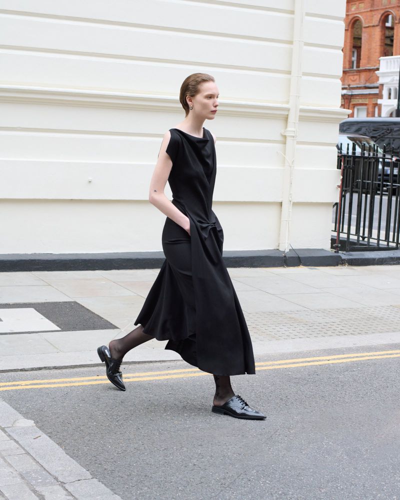 Woman in a black dress walking across a street with crosswalk markings in an urban environment with white building in background.