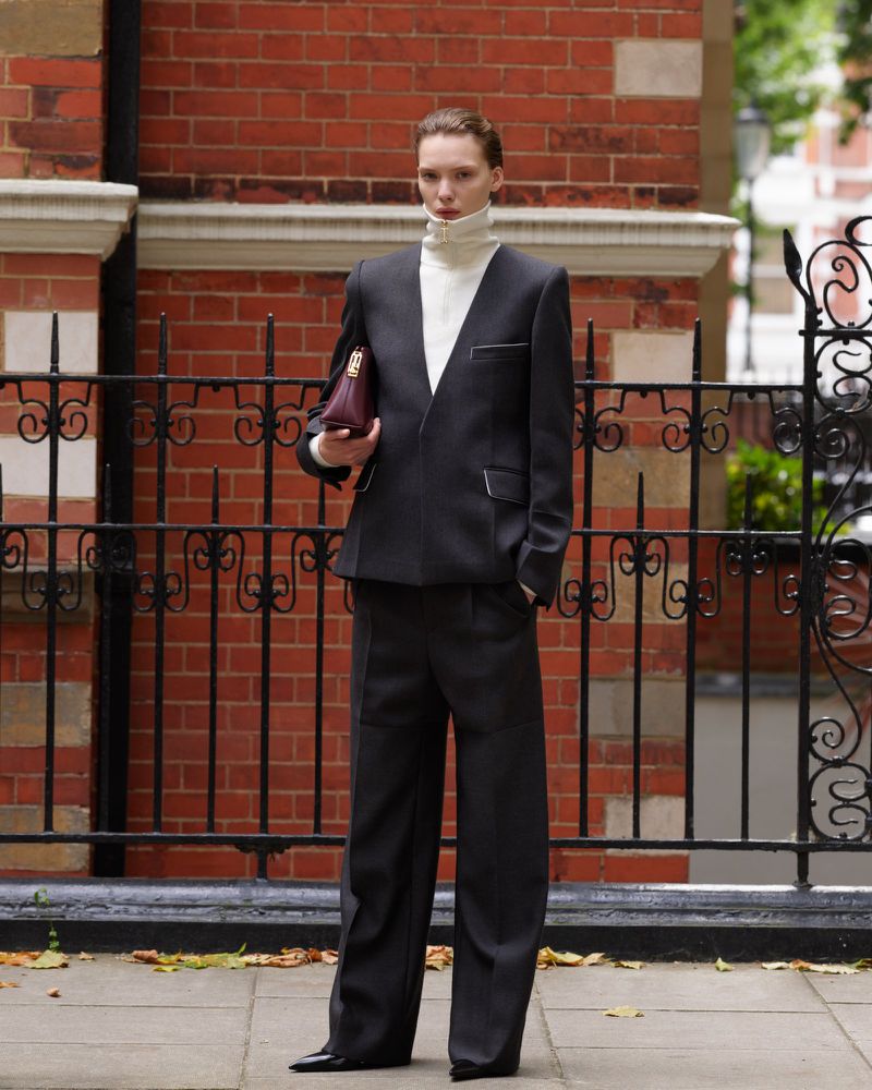 Elegant woman in a black suit and turtleneck, carrying a clutch, posing outdoors against a brick wall and wrought iron fence.