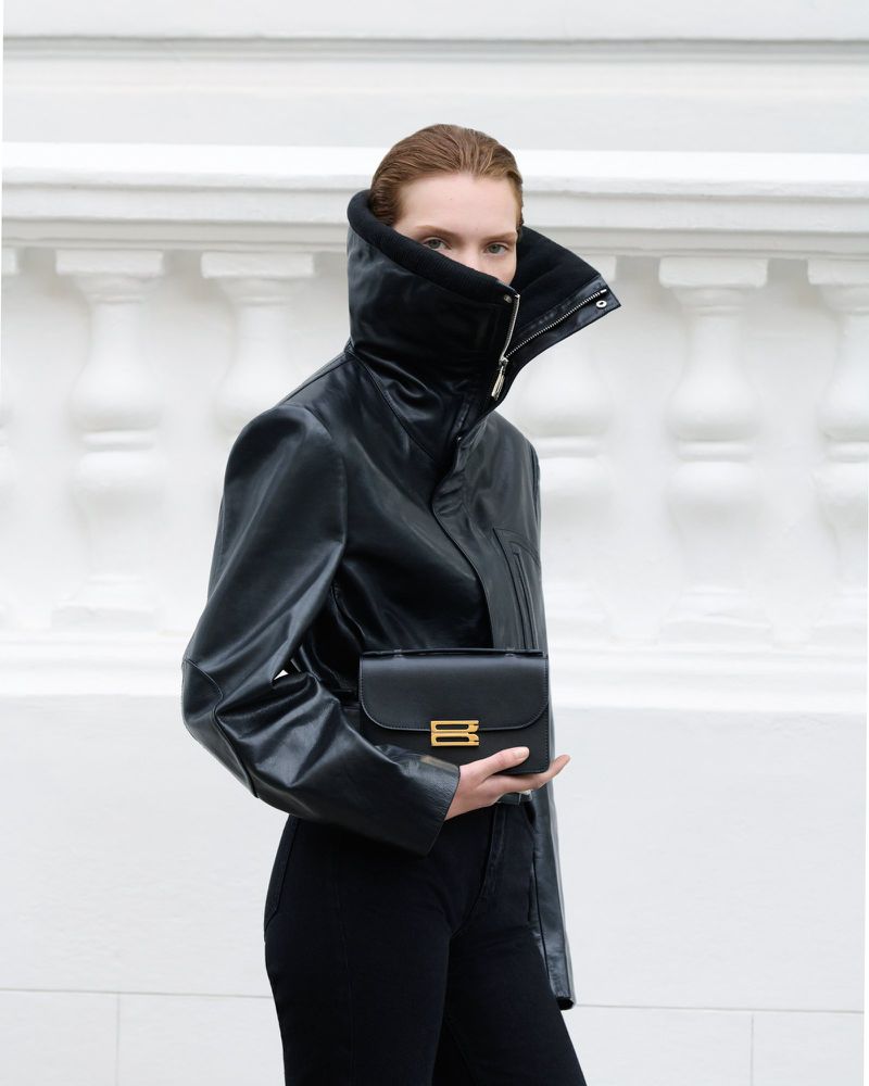 Fashionable woman in a black leather jacket and black pants holds a small black leather purse against a neutral white background.