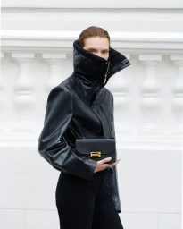 Model wearing a black leather jacket with a high zipped collar holding a black handbag against a white architectural background