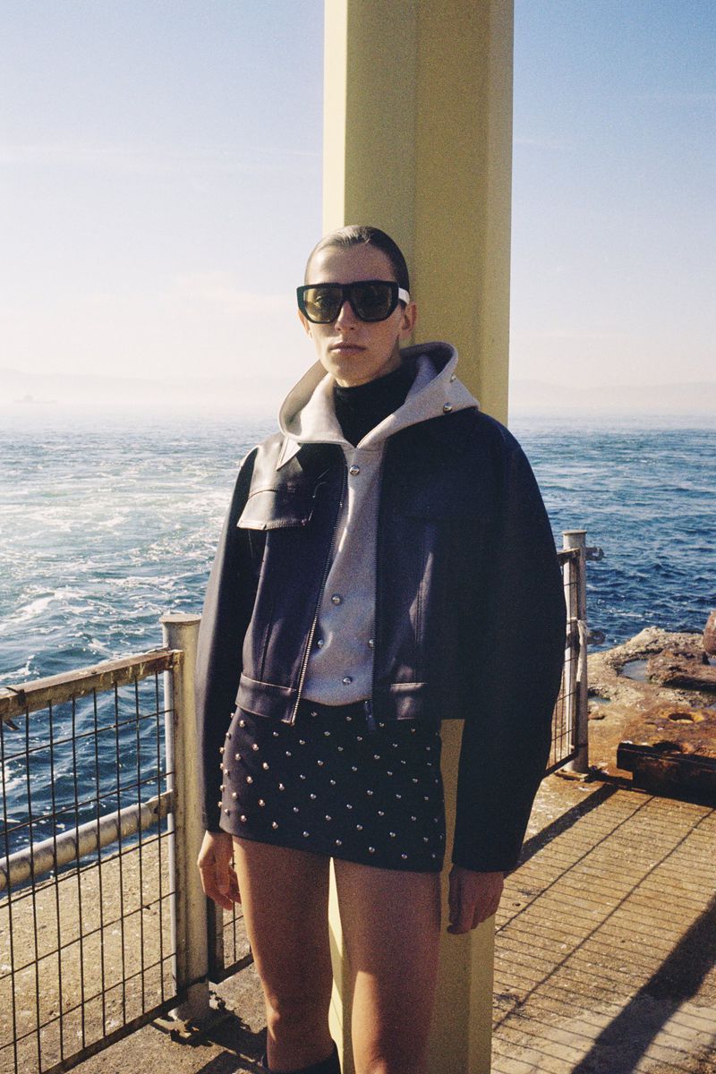 A person stands in a jacket, hooded shirt, and studded skirt on a seaside structure, wearing sunglasses under a bright sky.