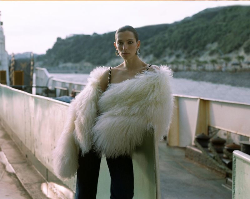Woman poses on a ferry in a cream fur coat, dark jeans, and statement earrings against a backdrop of water and a green tree-covered island.